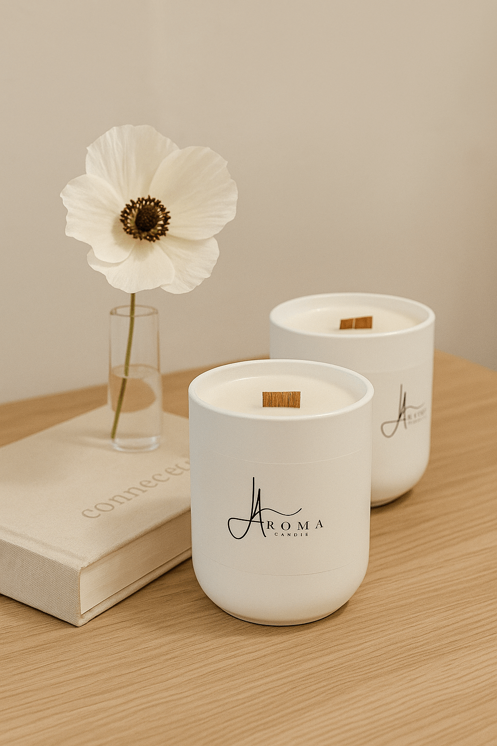 Two white candles with a brand logo on a wooden surface next to a white flower and book.